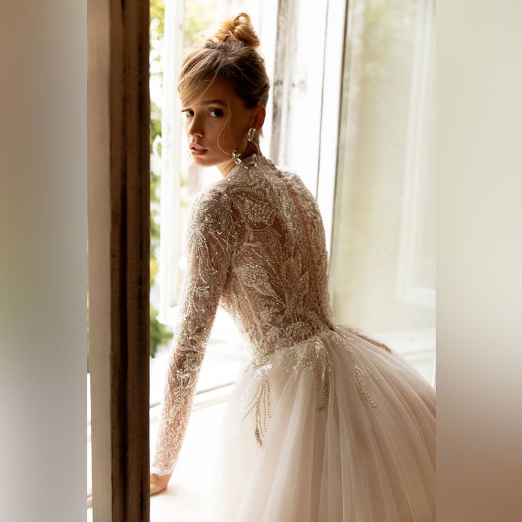 a woman in a wedding dress standing by a window looking at the camera with her hand on her hip