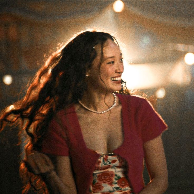 a woman with long hair smiling and wearing a red top in a dark room filled with lights