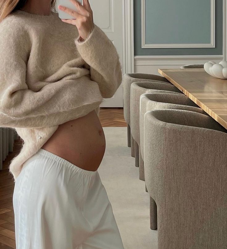 a pregnant woman taking a selfie in her living room while wearing white pants and a sweater