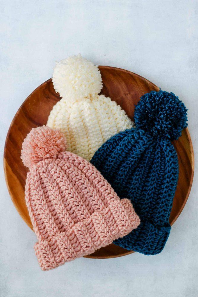 three crocheted beanies on a wooden plate with white, blue and pink yarn