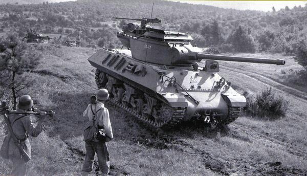 two men standing next to a tank in the grass with another man looking at it