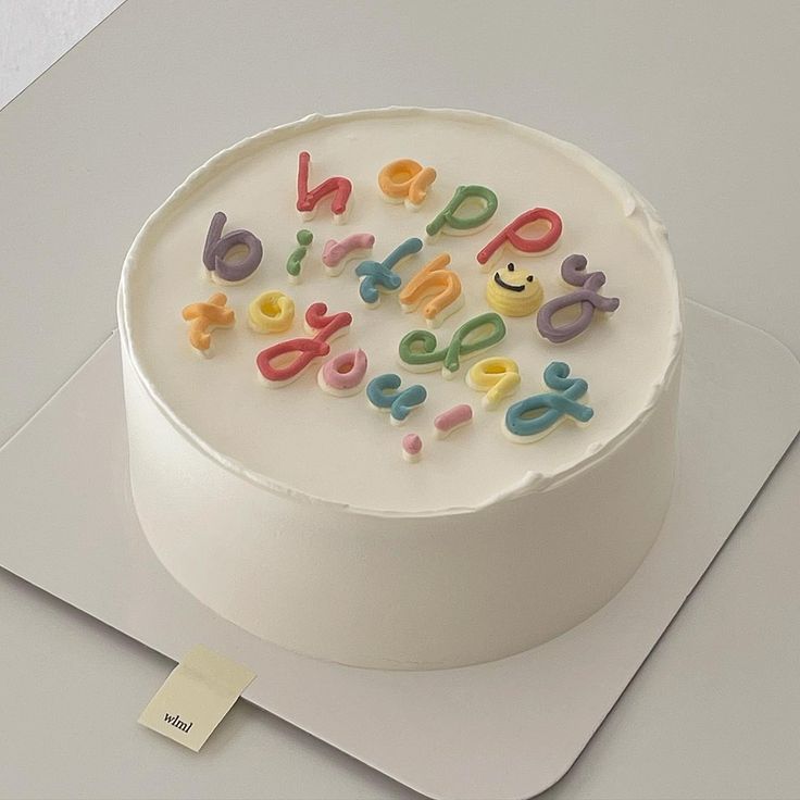 a white cake with colorful letters on it sitting on top of a table next to a piece of paper