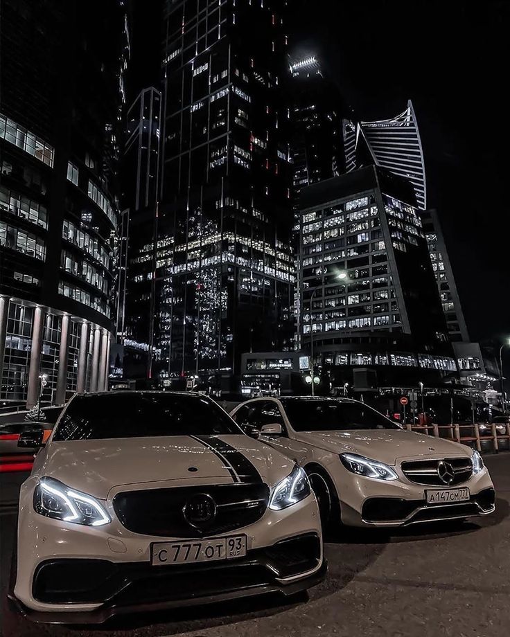 two mercedes benzs parked in front of tall buildings at night with city lights behind them