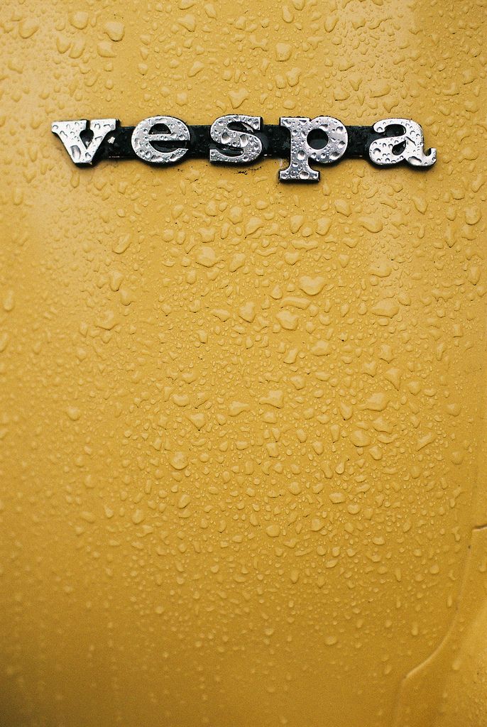 the word vespa is written in silver letters on a yellow car with drops of water