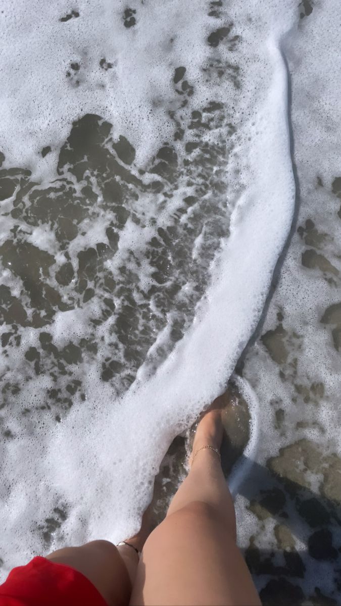 someone is standing in the water at the beach with their feet up on the sand