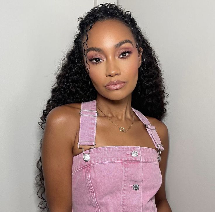 a woman with curly hair and overalls posing for a photo in front of a white wall