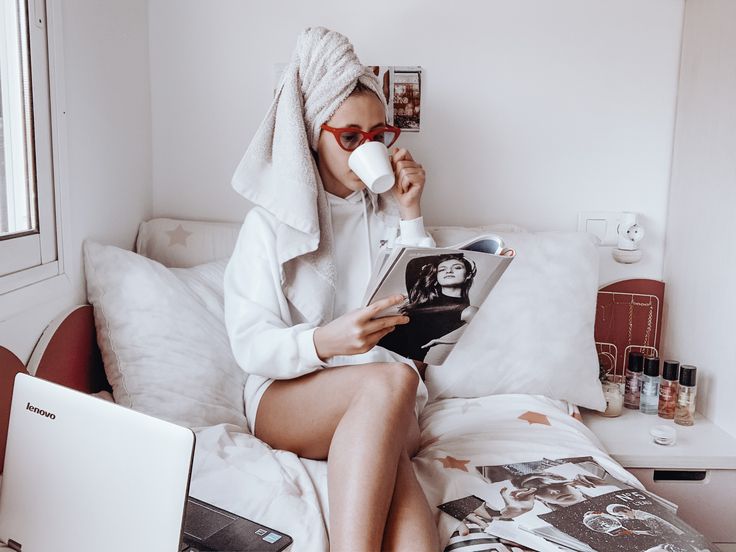 a woman sitting on a bed drinking from a cup while reading a book and using a laptop