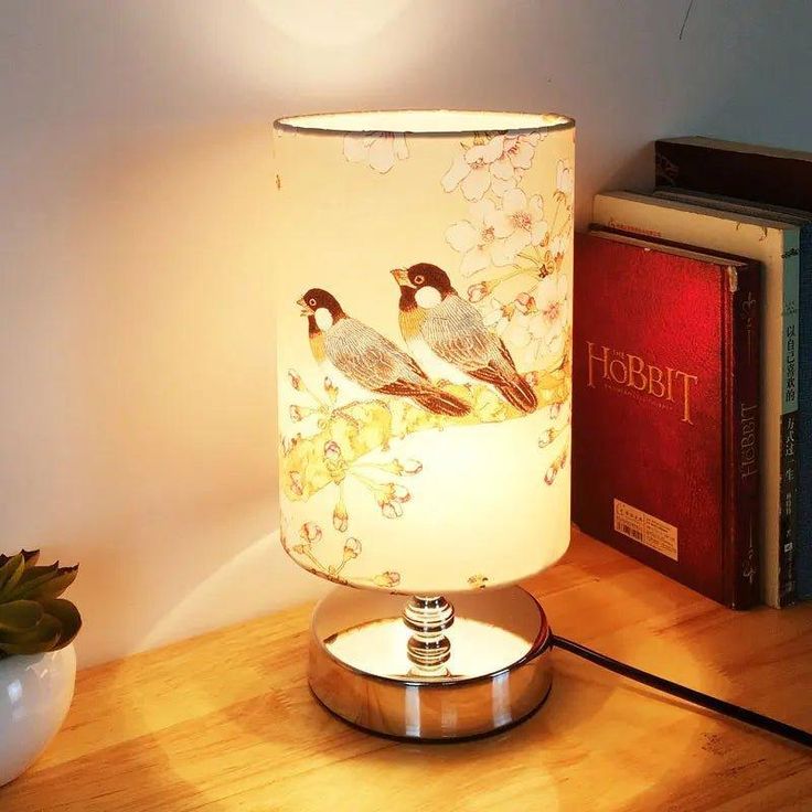 a lamp with two birds on it sitting next to a book and a potted plant