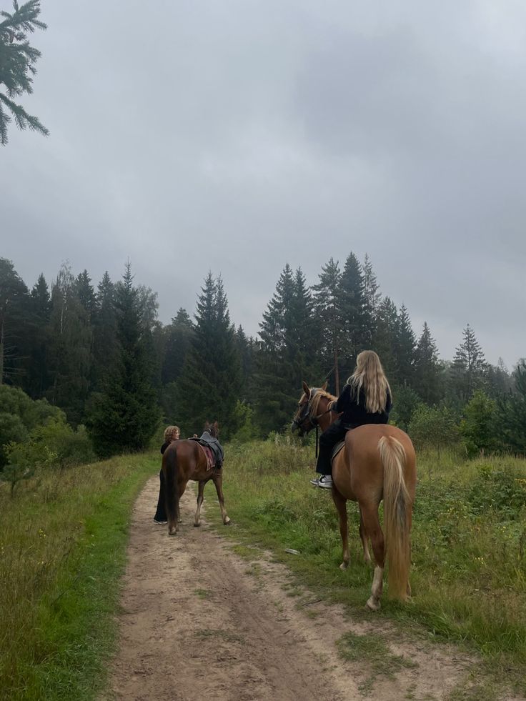 two people riding horses on a dirt road in the woods with trees and grass behind them