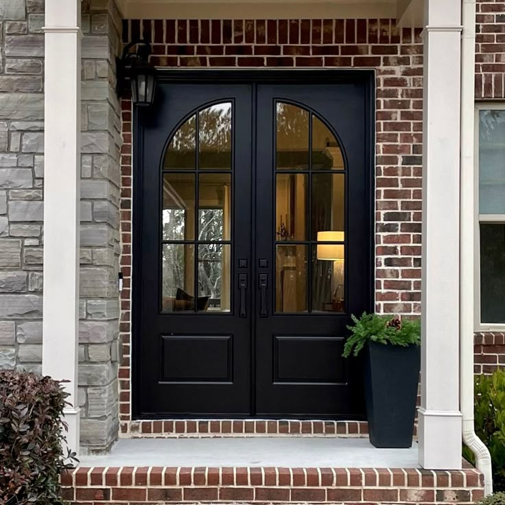 a black double door with two planters on the front steps and brick building behind it