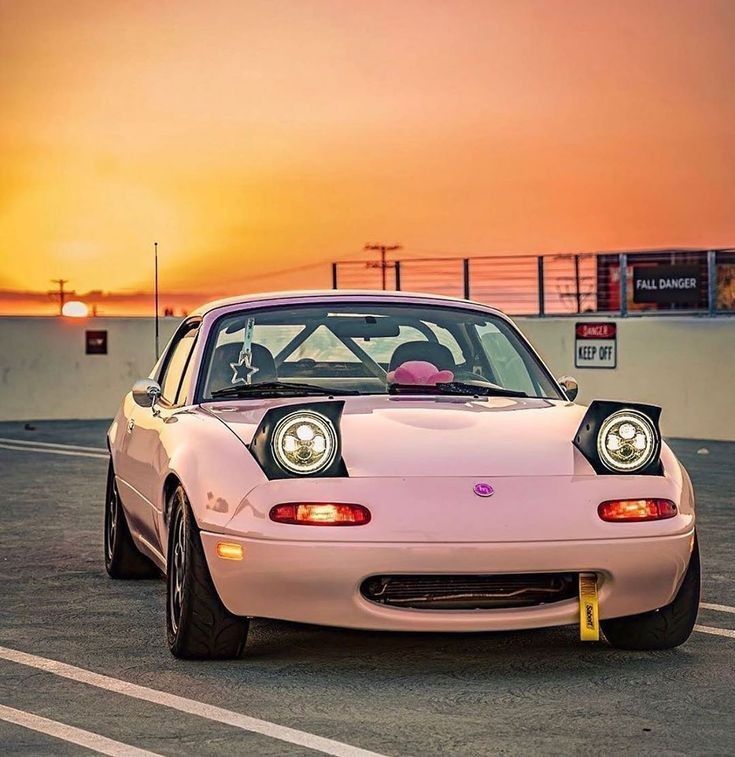 a pink car parked in a parking lot with the sun setting behind it's headlight