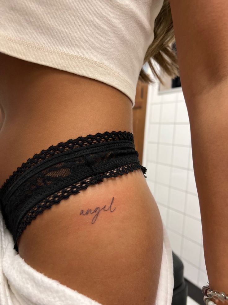 Angle Word Tattoos, Angel Tattoo On Hip, Women Tattoos Hip, Hip Tattoos Women Writing, Small Tattoo Areas For Women, Name On Hip Tattoo, Upper Thigh Word Tattoo, Angel Script Tattoo, Hip Small Tattoos Women