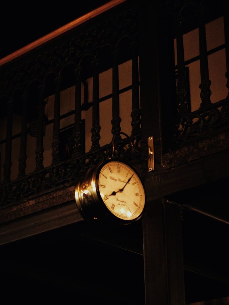 a clock that is on the side of a building in front of a balcony at night