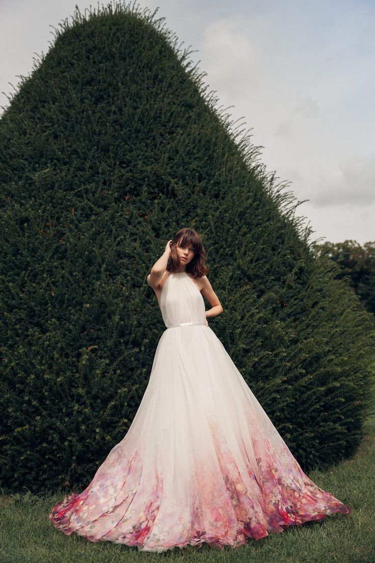 a woman standing in front of a tall bush wearing a white and pink wedding dress