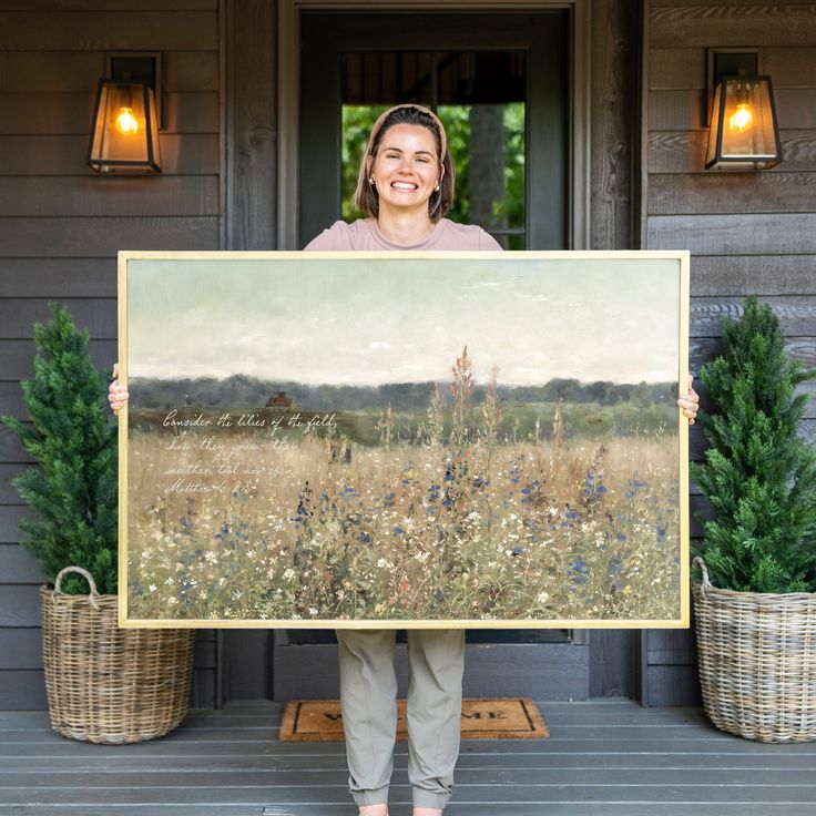 a woman holding up a large painting in front of a house with potted plants