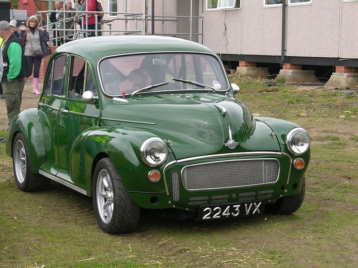 an old green car parked in front of a building with people standing around it on the grass