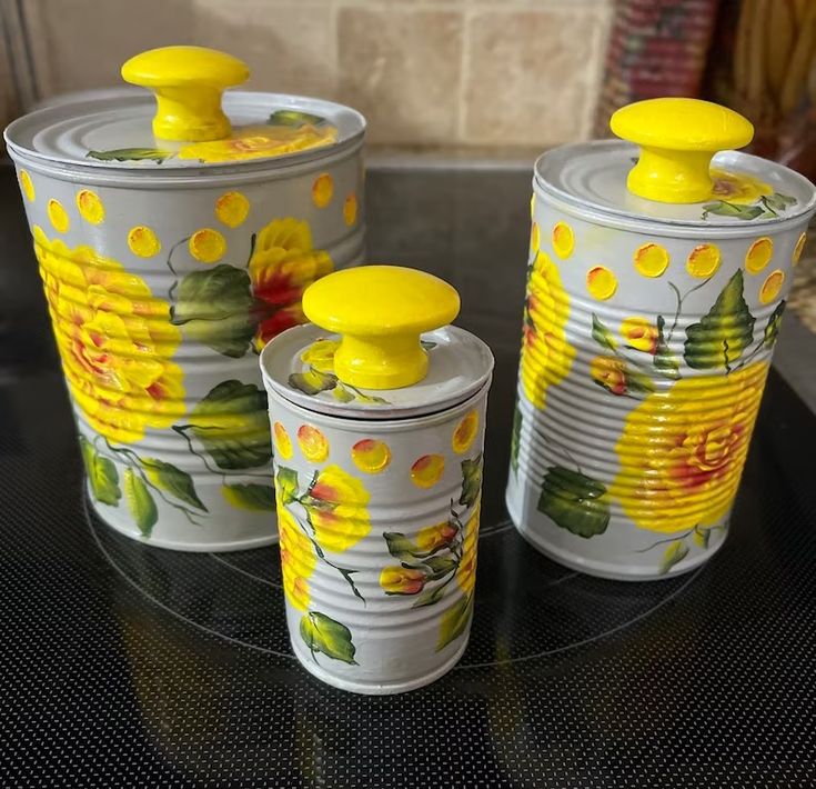 three tin canisters with yellow flowers painted on them sitting on a glass table