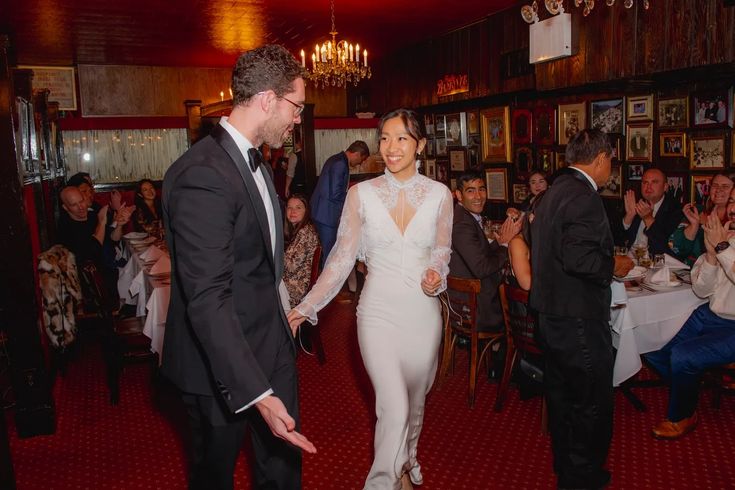 a man and woman in formal wear walking through a room with people seated at tables