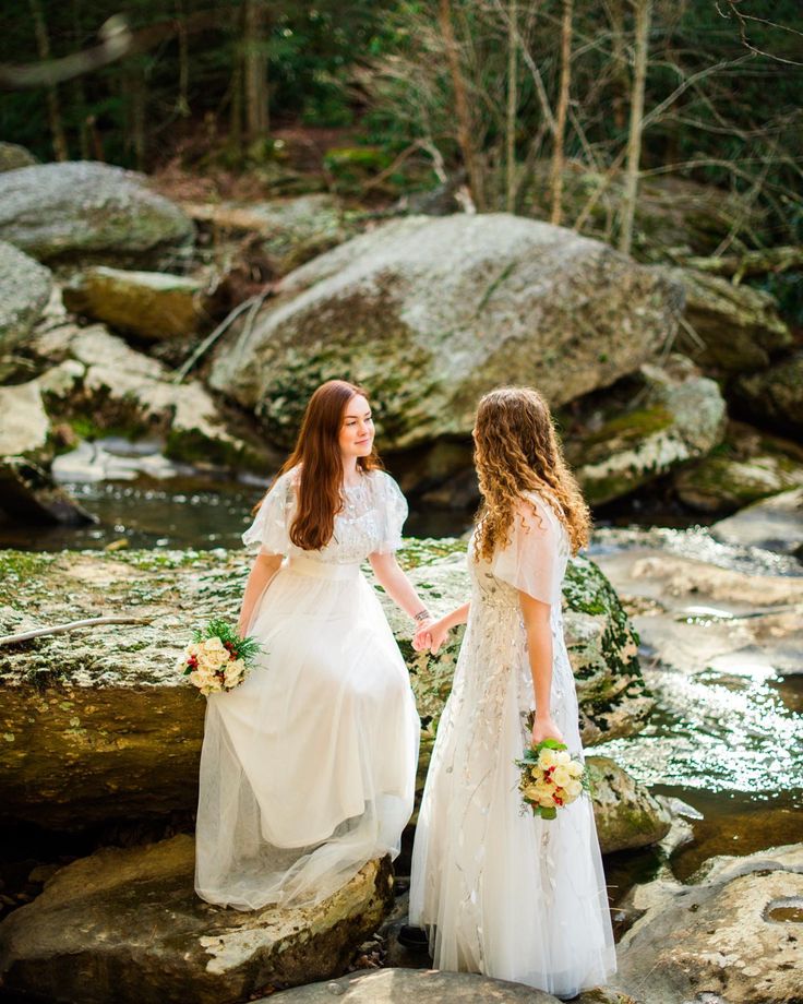 two brides standing on rocks in front of a stream and smiling at each other