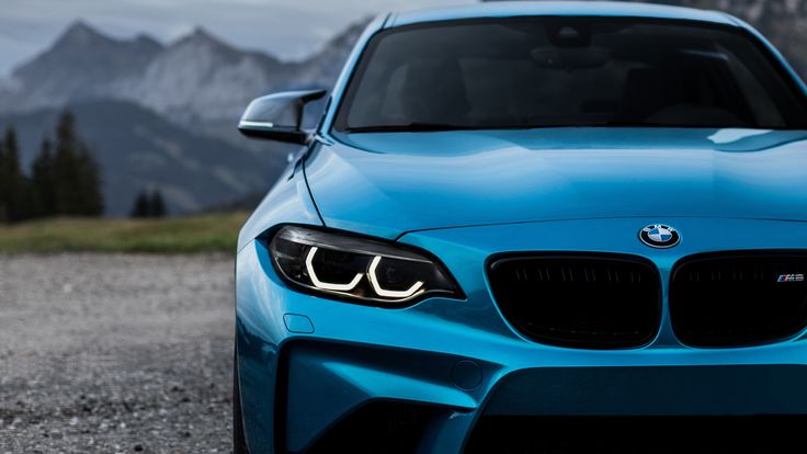 the front end of a blue bmw car parked on top of a gravel road with mountains in the background