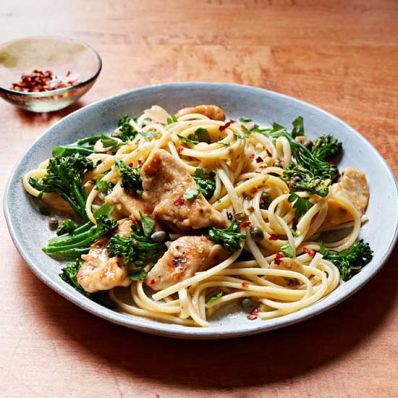 a plate of pasta with chicken and broccoli on a wooden table next to a glass of wine