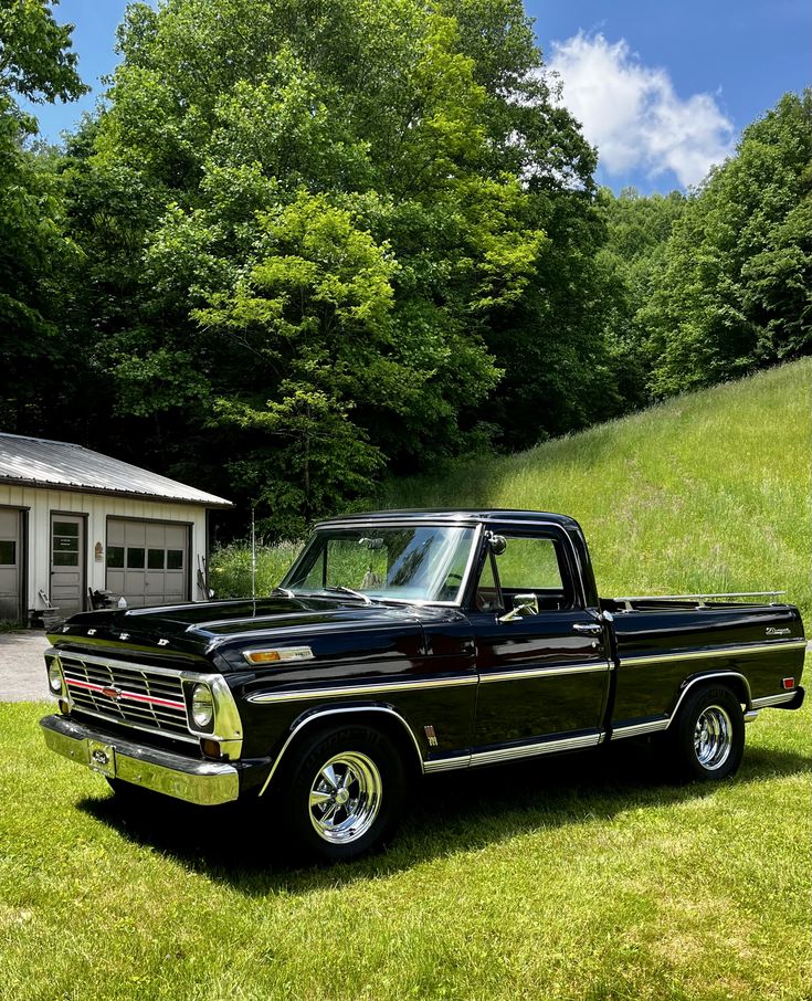 an old black pickup truck parked in front of a garage on a green field with trees