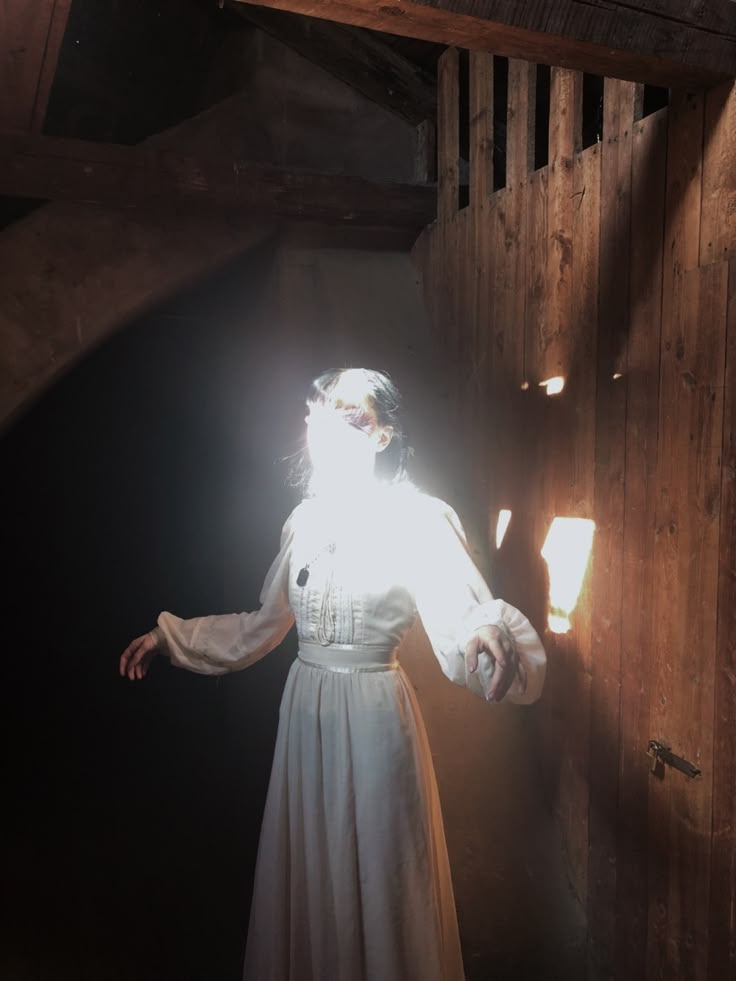 a woman in a white dress standing next to a wooden wall with sunlight coming through the window