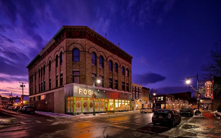 an empty street at night with cars parked on the side and buildings in the background