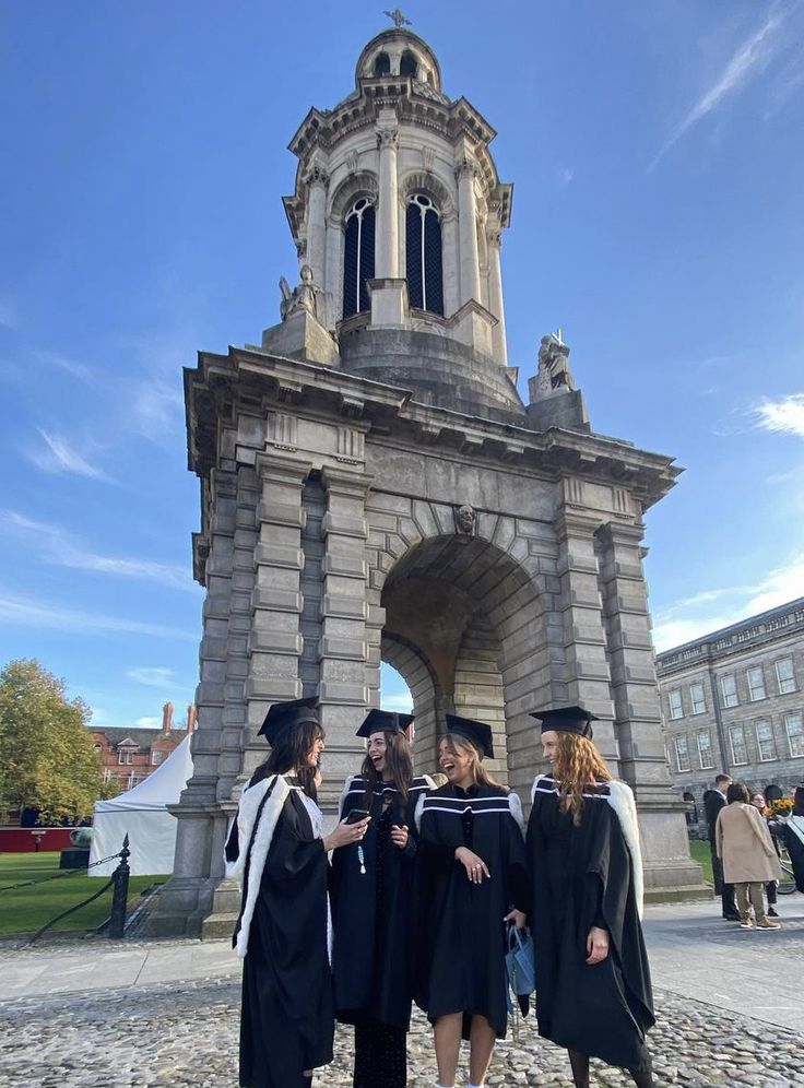 three women in graduation gowns are talking to each other near an old clock tower