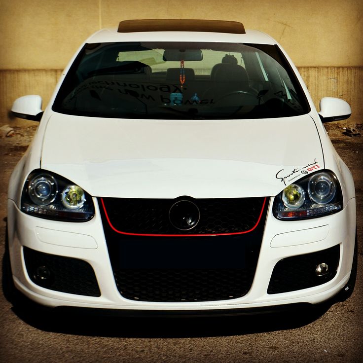 the front end of a white car parked in a parking lot next to a building