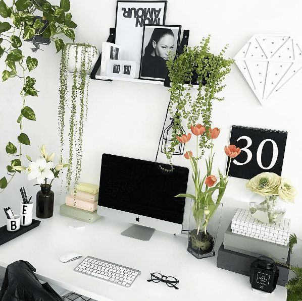 a white desk topped with a computer monitor next to a plant filled wall mounted clock