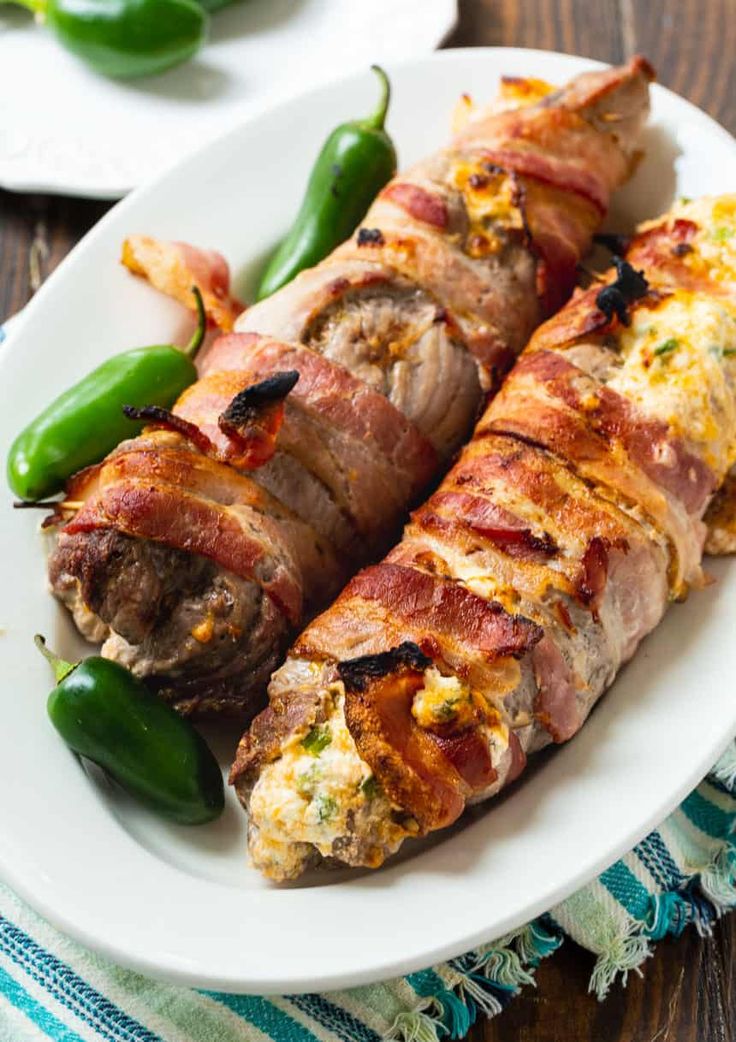 bacon wrapped stuffed peppers on a white plate with green peppers in the foreground and other food items to the side