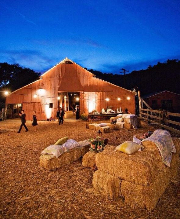 a barn with hay bales in the foreground and people walking by at night