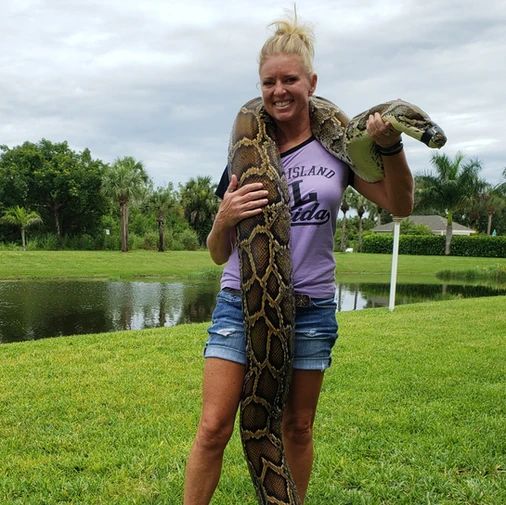 a woman is holding a large snake in her hands while standing on the grass near a pond