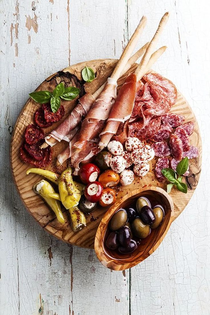 an assortment of meats and vegetables on a wooden platter with olives, tomatoes, peppers, bread