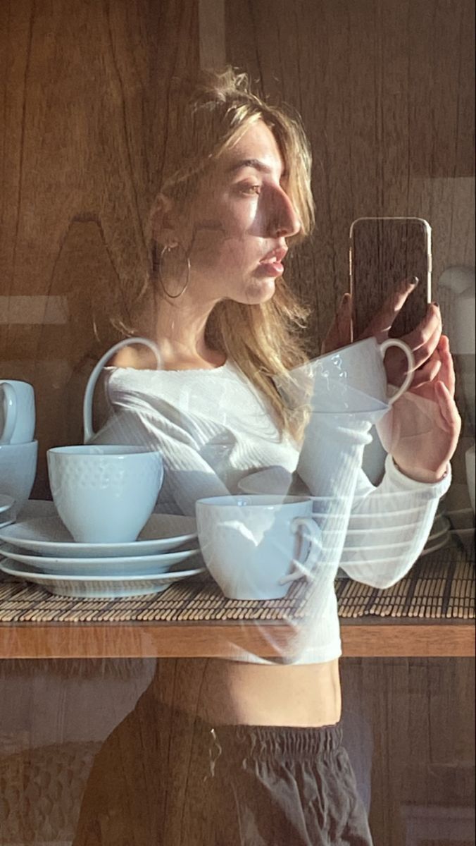 a woman taking a selfie with her cell phone in front of a shelf full of dishes and cups