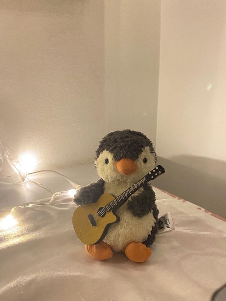 a stuffed penguin playing a guitar on a bed with lights in the room behind it