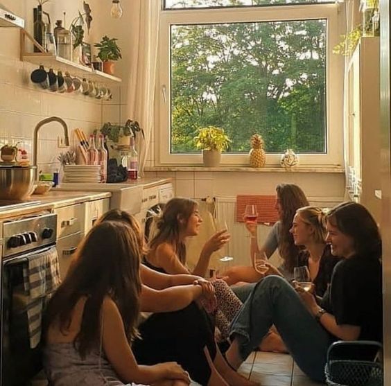 four women sitting on the floor drinking wine in a small kitchen with an open window