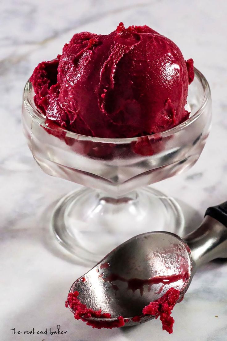 a scoop of red ice cream in a glass bowl with a spoon next to it