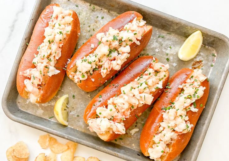 four hot dogs with toppings and chips on a baking sheet next to a lemon wedge