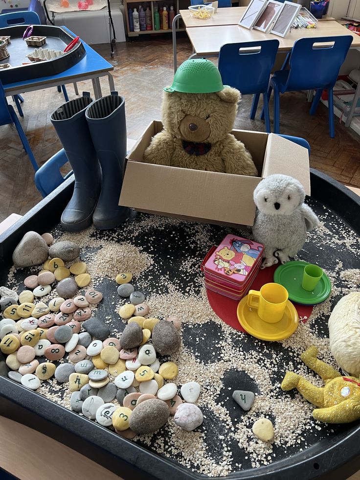 two teddy bears are sitting in a box filled with sand and rocks, while another bear is standing next to it