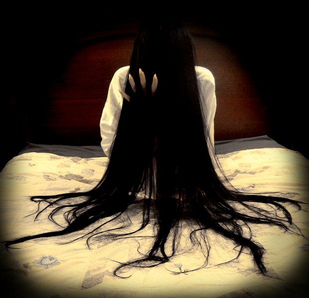 a woman with long hair sitting on a bed in the dark, looking down at her head