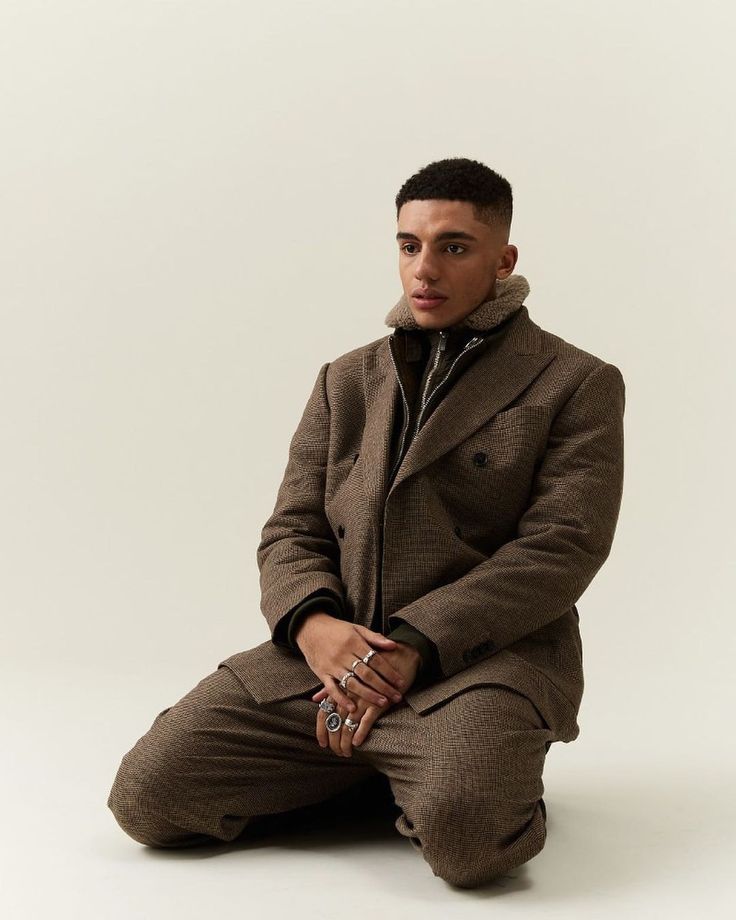 a man sitting on the ground wearing a brown suit and silver ring with his hands in his pockets