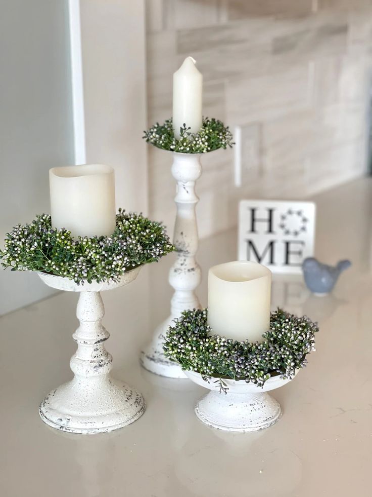 three white candles sitting on top of a table with greenery in each candle holder