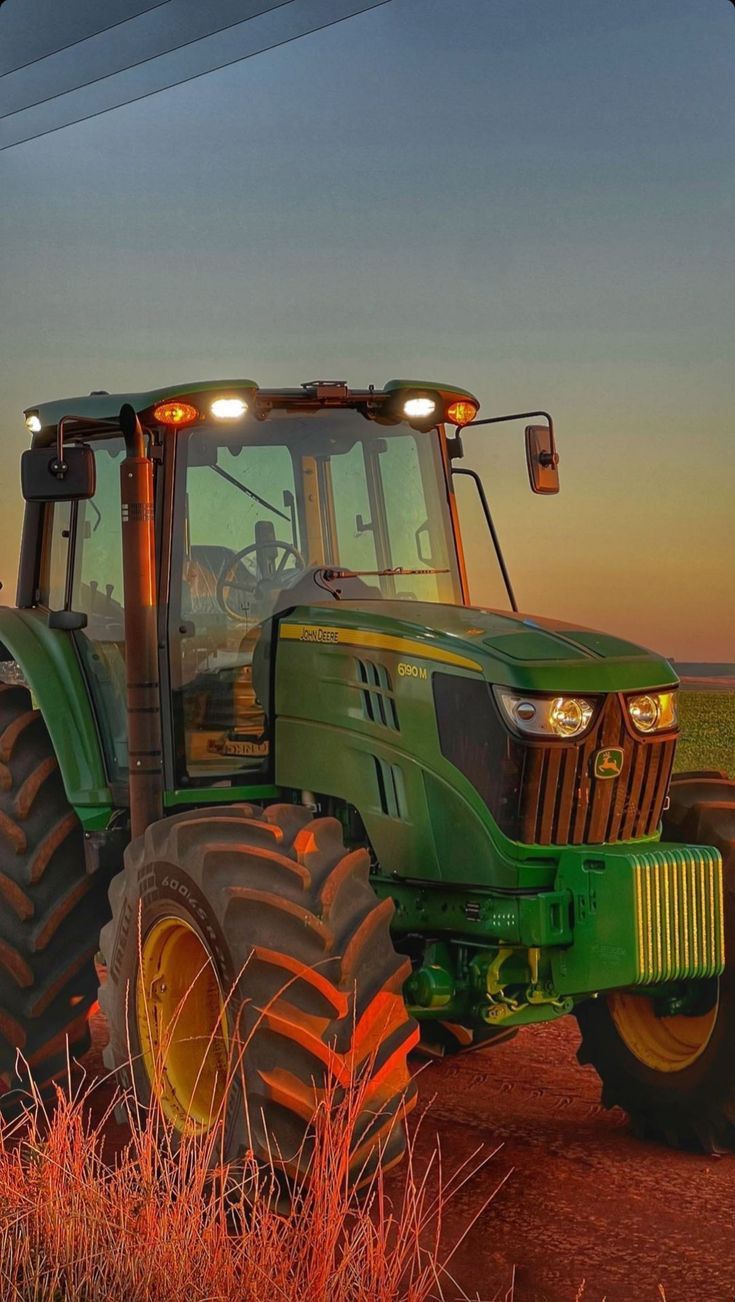 a green tractor parked in the middle of a field at sunset with its lights on