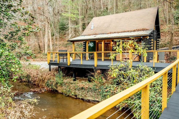 a small wooden cabin in the woods next to a river with a bridge over it