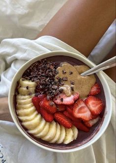 a person is holding a bowl of fruit with chocolate and strawberries in it while sitting on a bed