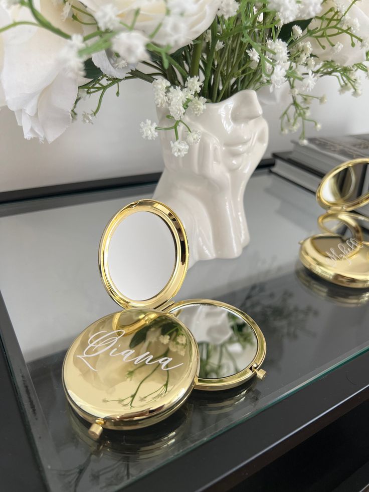 two gold compact mirrors sitting on top of a table next to a vase with white flowers