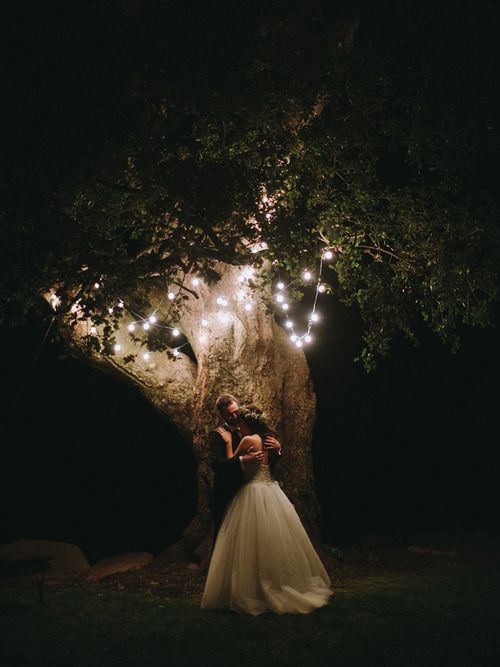 a bride and groom standing in front of a tree with fairy lights strung all over it