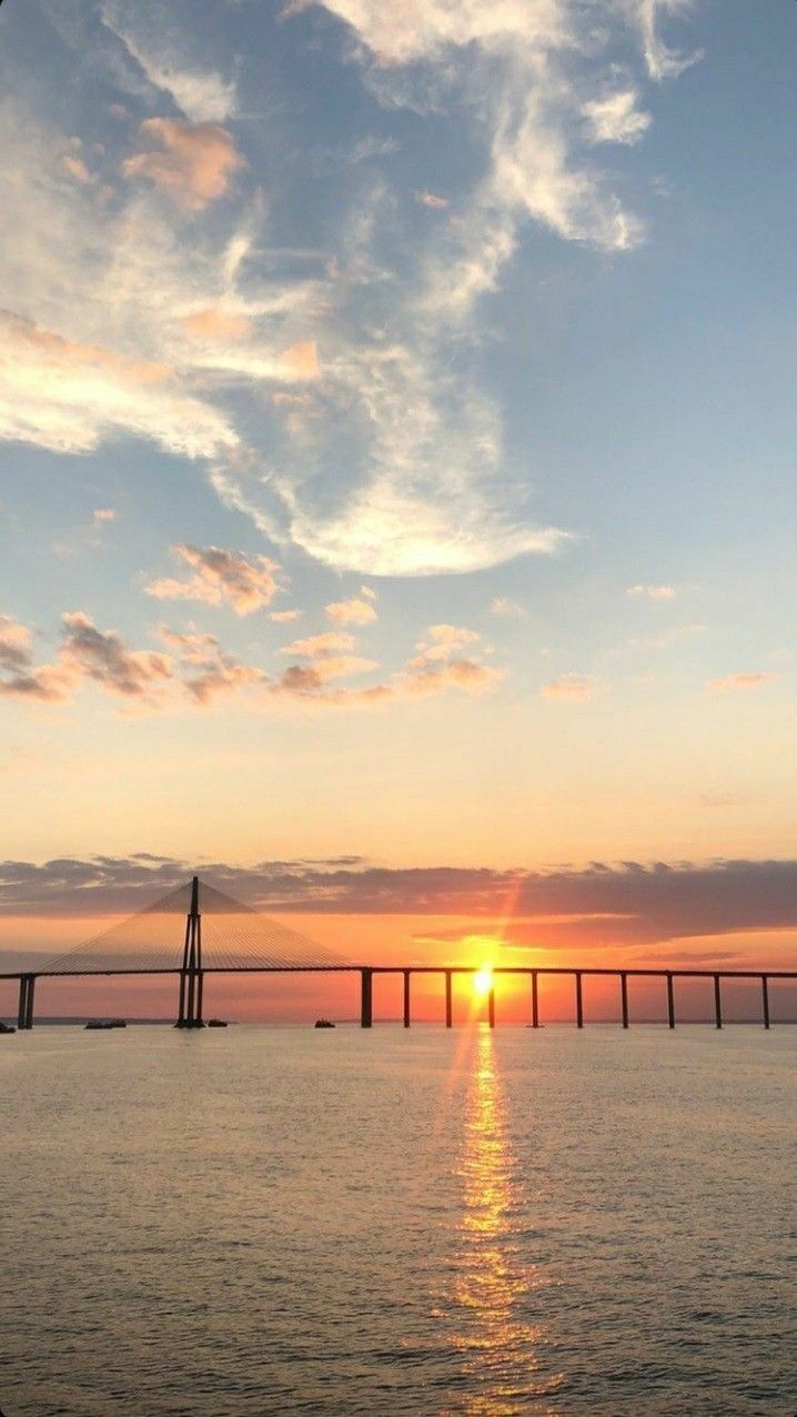 the sun is setting over an ocean with a large bridge in the distance and some clouds